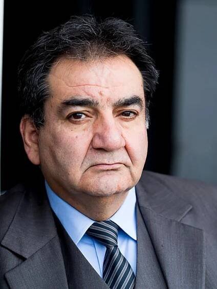 Mohammad-Ali Behboudi trägt einen grauen Anzug und Krawatte, lehnt an einer Wand und schaut ernst in die Kamera.