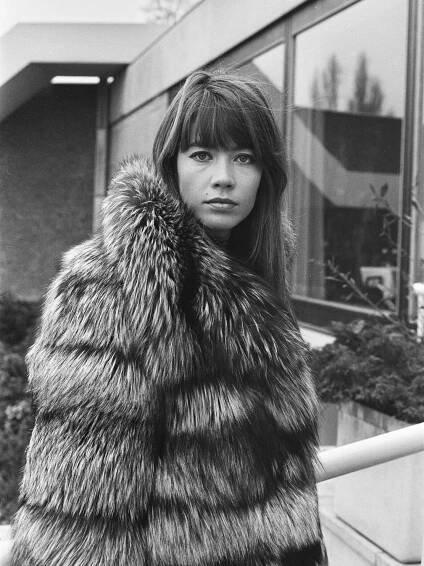 A close-up black-and-white photo of her wearing a fur coat and looking directly at the camera outside a building with a tall glass window. She had long hair and bangs.