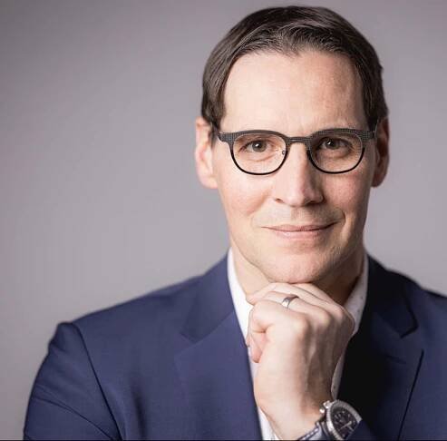 Jürgen Müller, Brille und blaues Jackett, stützt das Kinn auf die Hand und blickt vor grauem Hintergrund nachdenklich in die Kamera.