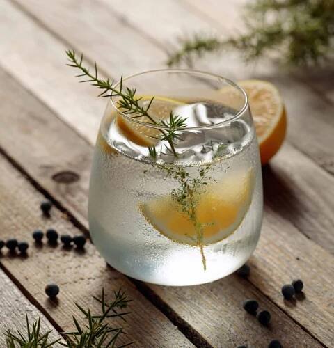 A chilled gin and tonic with lemon and rosemary in a round glass on a wooden table, with juniper berries scattered around.