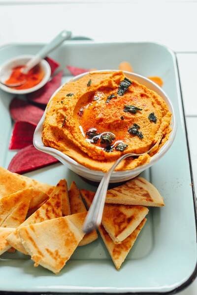 Close up shot of a bowl of White Bean Pumpkin Hummus on a tray with pita slices, beet, and carrot