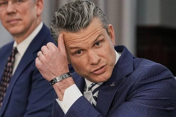 Pete Hegseth scratches the right of his forehead near his hairline with his left thumb as he sits at a table near a man.