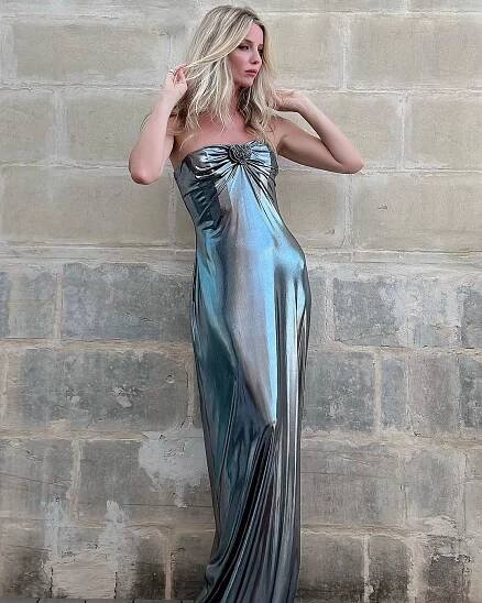 A woman in a sleek, metallic blue gown stands against a beige stone wall, her fingers running through her blonde hair. The dress features an Annabelle Wallis-inspired bow detail at the neckline.