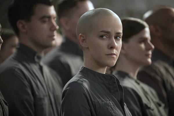 Person with a shaved head in a uniform, looking to the side, standing among others in similar attire; scene suggests a serious or dystopian setting