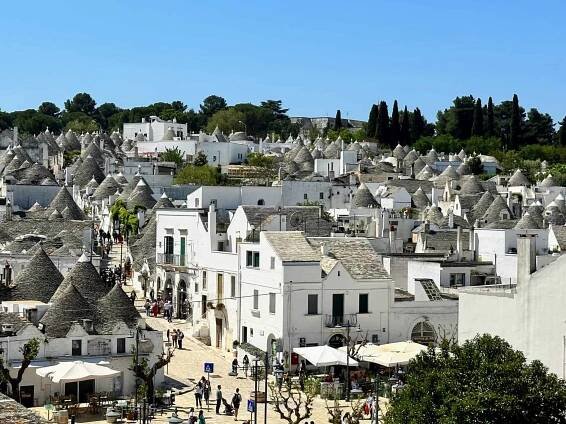 View of the trulli and white buildings of Rione Monti in Alberobello, Italy. You can also see people walking in the piazza and streets.
