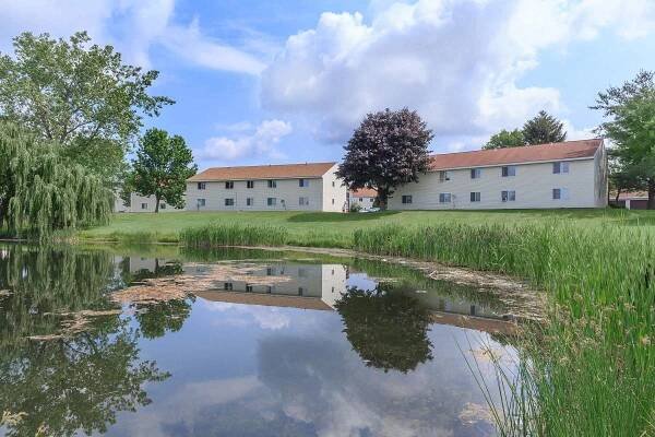 A serene landscape featuring two buildings with light-colored exteriors and red roofs, situated next to a pond. The pond reflects the buildings and surrounding greenery, including grassy areas and trees. Fluffy white clouds drift across a bright blue sky.