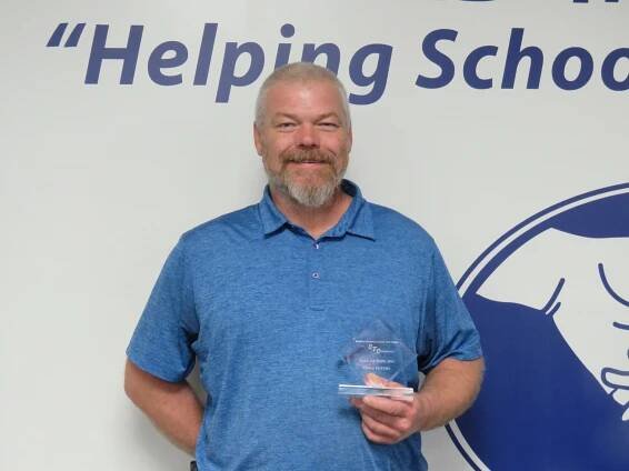 A man proudly holds an award in front of a wall displaying the phrase "Helping Schools."