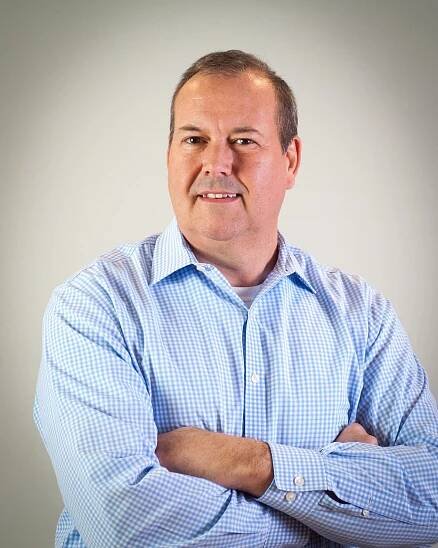 A man in a blue shirt is standing with his arms crossed and smiling.