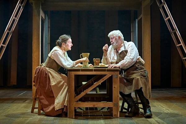 Liza Sadovy (Mary), wearing a white shirt, green corset and skirt in shades of brown, sits at a wooden table. Opposite her sits Peter Wight (John) with white hair and white beard, wearing a black waistcoat and brown apron.  Photo by Manuel Harlan © RSC
