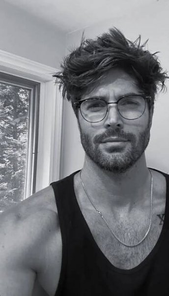 a man wearing glasses and a black tank top is posing for a photo in front of a window