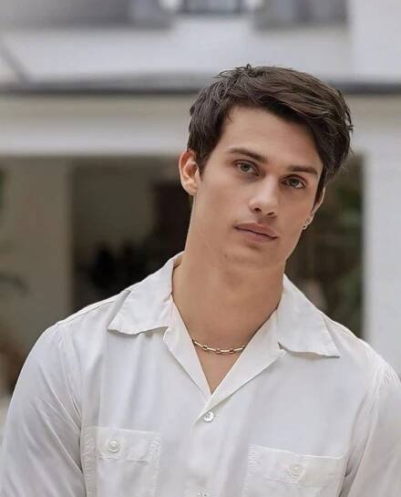 a young man wearing a white shirt looks at the camera while standing in front of a house
