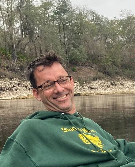 Matthew Carrigan, wearing glasses and a green hoodie, smiles while looking away from the camera in front of a body of water and trees.