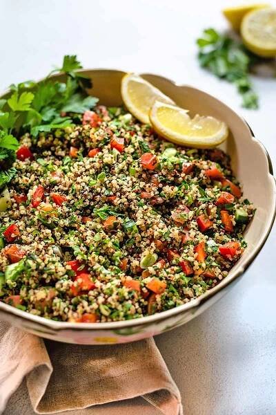 Closely cropped oblique vertical shot of a large, beige, ceramic serving bowl of quinoa tabbouleh with fresh herbs and lemon, on a folded beige cloth on a white and gray speckled surface, with more citrus and herbs in soft focus in the background.