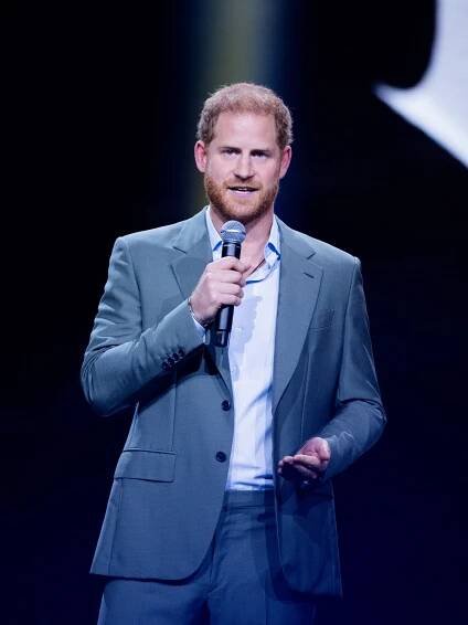 Prince Harry wears a blue suit on stage and holds a microphone