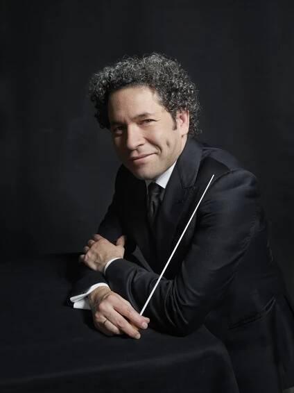 Gustavo Dudamel mit lockigem Haar im Anzug lehnt an einem dunklen Tisch und hält einen Taktstock vor einem dunklen Hintergrund. Er hat Grübchen und blickt direkt in die Kamera.