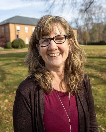 A woman with wavy, light brown hair, wearing glasses, a maroon top, and a dark cardigan, smiles outdoors in a grassy area with a brick building and trees in the background.