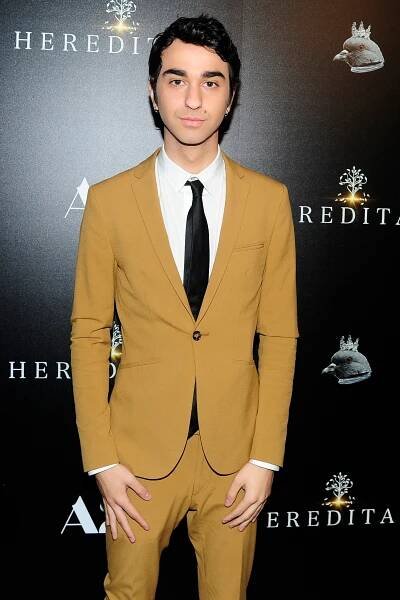 Person in a fitted suit with white shirt and black tie on a red carpet, standing against a backdrop with "Hereditary" logos