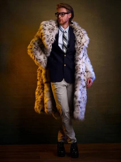 Person in glasses models stylish navy blazer and long spotted fur coat with khaki pants against dark backdrop.