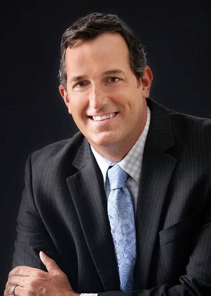Professional man in a dark suit and light blue tie smiles confidently against a dark background during his personal branding portrait session with Mueller Photography in La Crosse, WI.