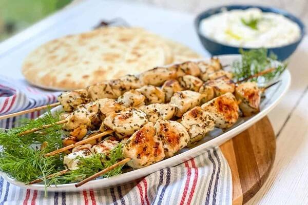 Side view of chicken souvlaki skewers on a white platter with pita and tzatziki sauce