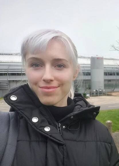 Selfie image of Esther González-Almela, wearing a black turtleneck sweater under a black winter jacket, in front of a building.