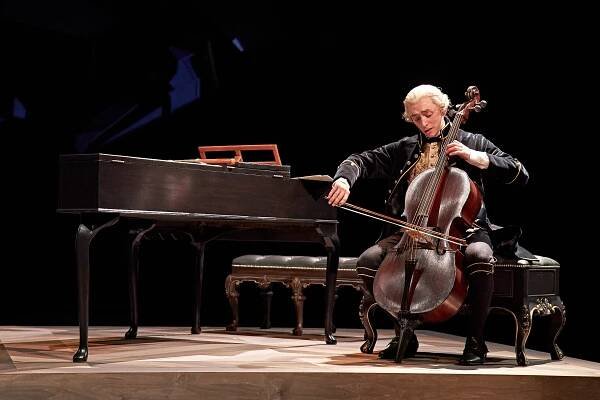 Samuel Blenkin is sat next to a Harpsicord playing a cello.