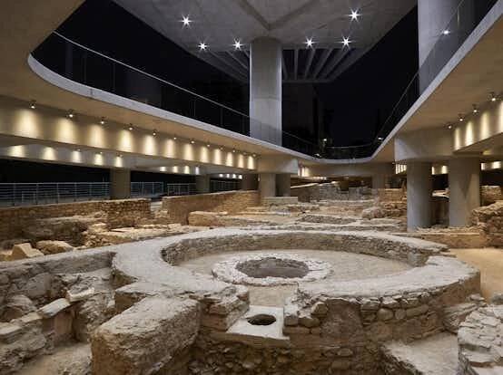 Archeologické zříceniny osvětlené moderním stropním osvětlením s kruhovou strukturou v popředí a vyvýšenými chodníky nad nimi.