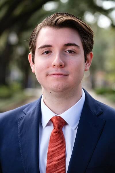 Professional headshot of a young professional with brown hair in a blue suit, with a white shirt and an orange tie.