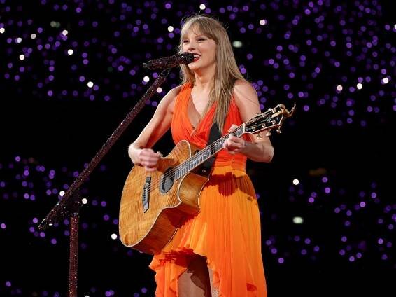 Taylor Swift smiles as she plays guitar on stage in an orange dress