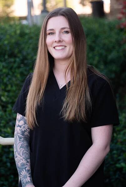 A woman with long hair and a tattoo on her arm is wearing a black scrub top.