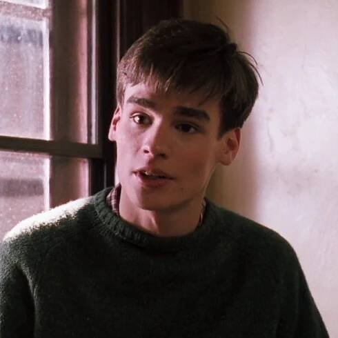 a young man standing in front of a window wearing a sweater and looking at the camera