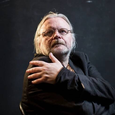 The writer Jon Fosse sits in a chair with an arm around himself. He is wearing glasses a black jacket and a scarf.