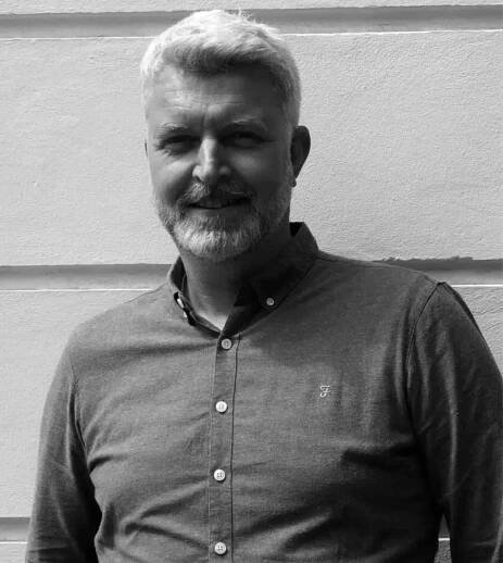 Man with a beard and short hair stands in front of a light-colored wall, wearing a button-up shirt. Black and white photo.