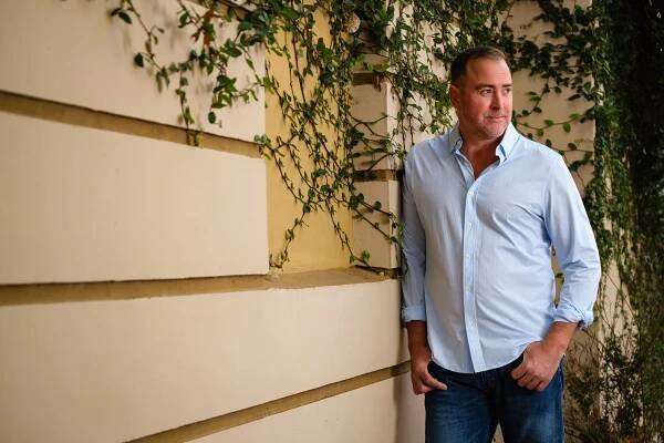 A smiling man wearing a blue shirt and jeans leans against a wall of ivy.