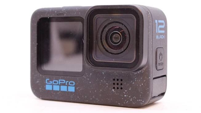 A close-up of a GoPro Hero 12 Black action camera. The lens is on the right, and a small screen is on the left. The camera is black with blue speckled details and model branding on the front.