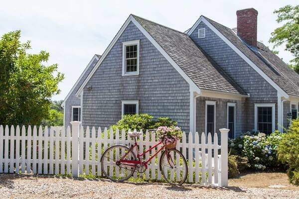 A weathered gray shingle house with white trim sits behind a white picket fence. A pink bicycle with a basket of flowers is leaning against the fence, and greenery surrounds the house on a sunny day.