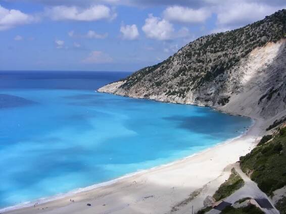 A view of Myrtos Beach in Kefalonia from above, showing turquoise waters meeting a white sandy shore. A steep, rocky hillside covered in green vegetation rises along one side of the beach. A winding road leads down to the shore, with a few people visible on the sand.