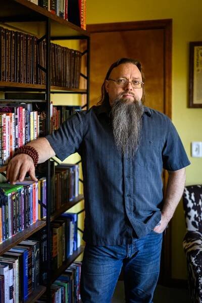 David Carson wears a dark blue short-sleeve shirt and jeans and has a long beard. He rests one arm on a bookshelf and the other hand in his pocket.