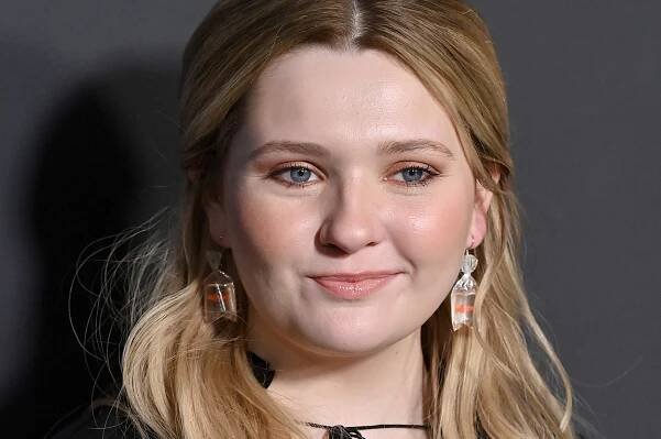 Abigail Breslin with long hair in a black dress poses for a photo. They are wearing earrings and have a slight smile