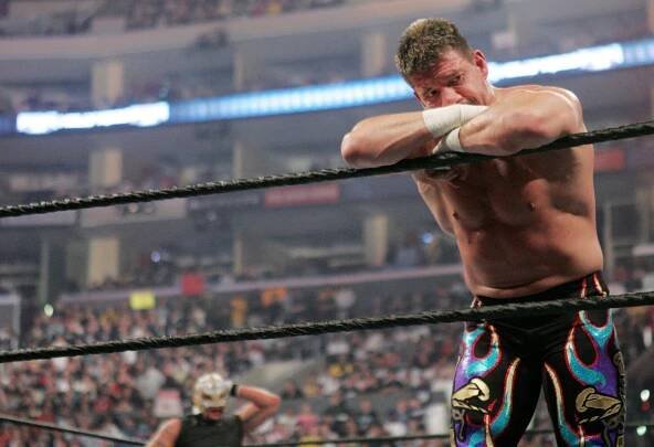 Eddie Guerrero, looking muscular and sweaty with no shirt, leans against the ropes of a ring. His opponent and an arena full of people watch.