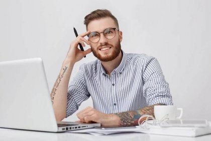 Free photo picture of thoughtful tattooed male enterpreneur with thick beard and trendy hairstyle begins work during morning