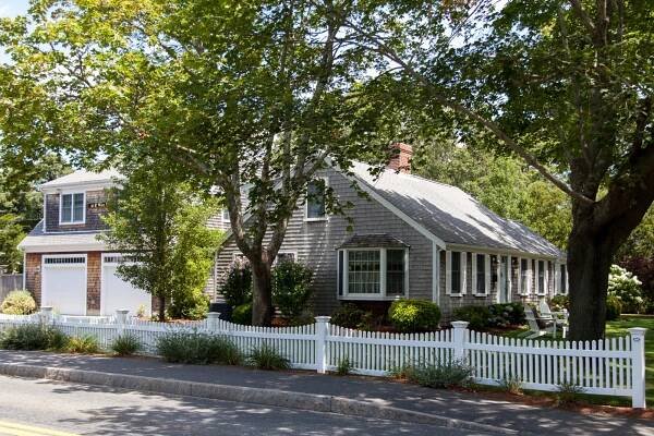 A grey shingled house with a white picket fence and a detached garage sits behind leafy trees on a sunny day. There is a garden and shrubs along the fence, with a road in the foreground.