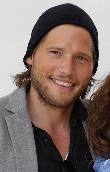 a man with long hair wearing a black beanie smiles at the camera while standing next to a woman