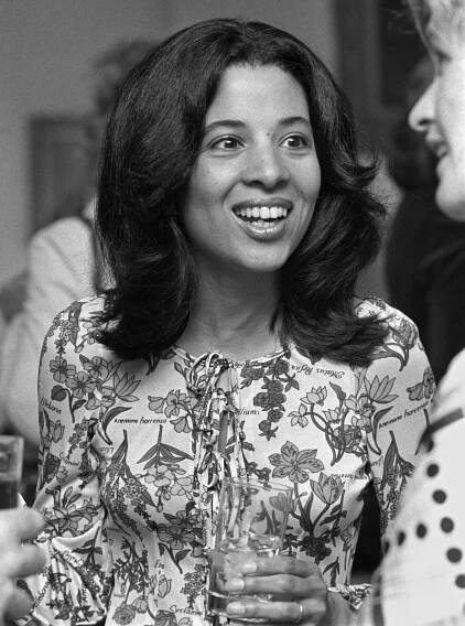 Gail Lumet Buckley, a young woman wearing a blouse that features flowers and botanical names and holding a drink in her hand, talks to people at a party.