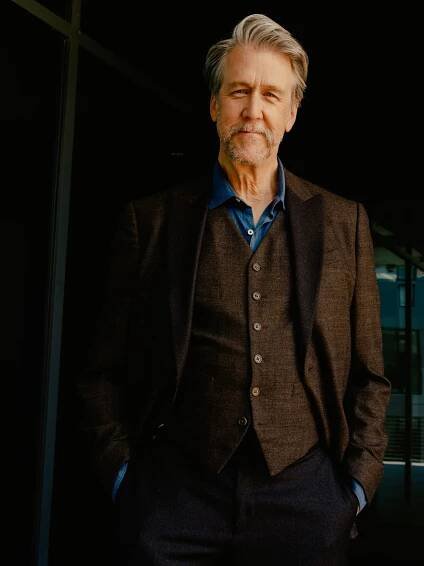 A man in a brown three piece suit and blue shirt smiles at the camera.