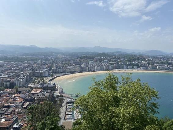A panoramic view of San Sebastián, Spain, showcasing the stunning La Concha Bay with its golden sandy beach curving along the turquoise waters. The cityscape features a mix of historic and modern architecture, with densely packed buildings leading to green hills in the background. Boats are docked in the marina, and lush trees frame the foreground, adding depth to the coastal scenery.