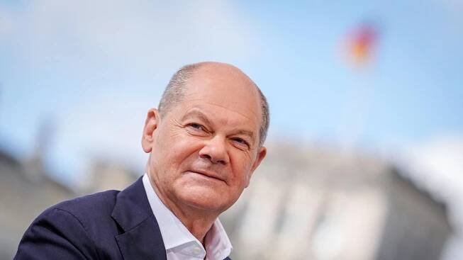 Bundeskanzler Olaf Scholz (SPD) lächelt in die Kamera. Im Hintergrund ist verschwommen der Berliner Reichstag zu sehen mit der Deutschlandflagge auf dem Dach. 