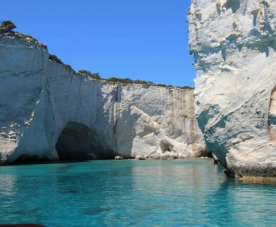 A scenic view of a tranquil body of water with a cave in the background, surrounded by cliffs and mountains, with Milos island visible in the distance under a clear sky.