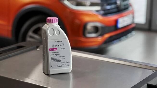Flasche Volkswagen Original Kühlmittelzusatz G12evo auf einem Rollcontainer, im Hintergrund ein VW T-Cross