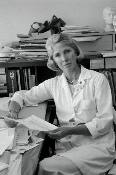 A black and white photo of Frances Conley wearing a white medical coat and sitting at a cluttered desk, a paper in her hand, her head tilted as she looks at the camera.